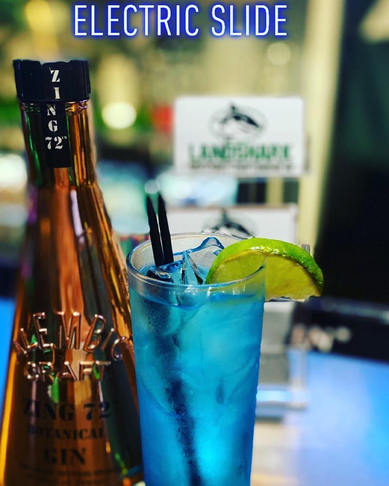 Blue cocktail with lime on a bar, labeled “Electric Slide,” beside a copper tap handle.