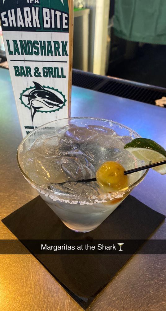 Cocktail in a stemmed glass with a lime wedge and cherry on a bar coaster, Shark Bite bar sign behind it