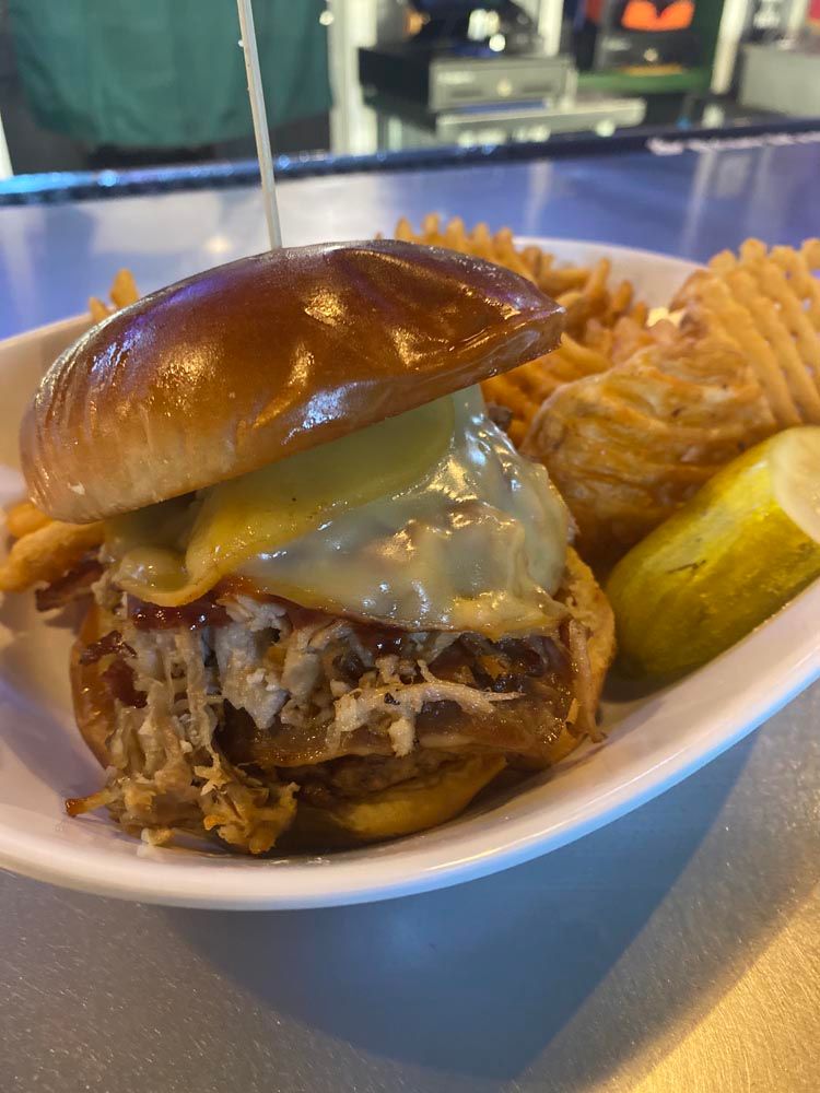 Pulled pork cheeseburger with chips and pickles on a white plate at a restaurant table
