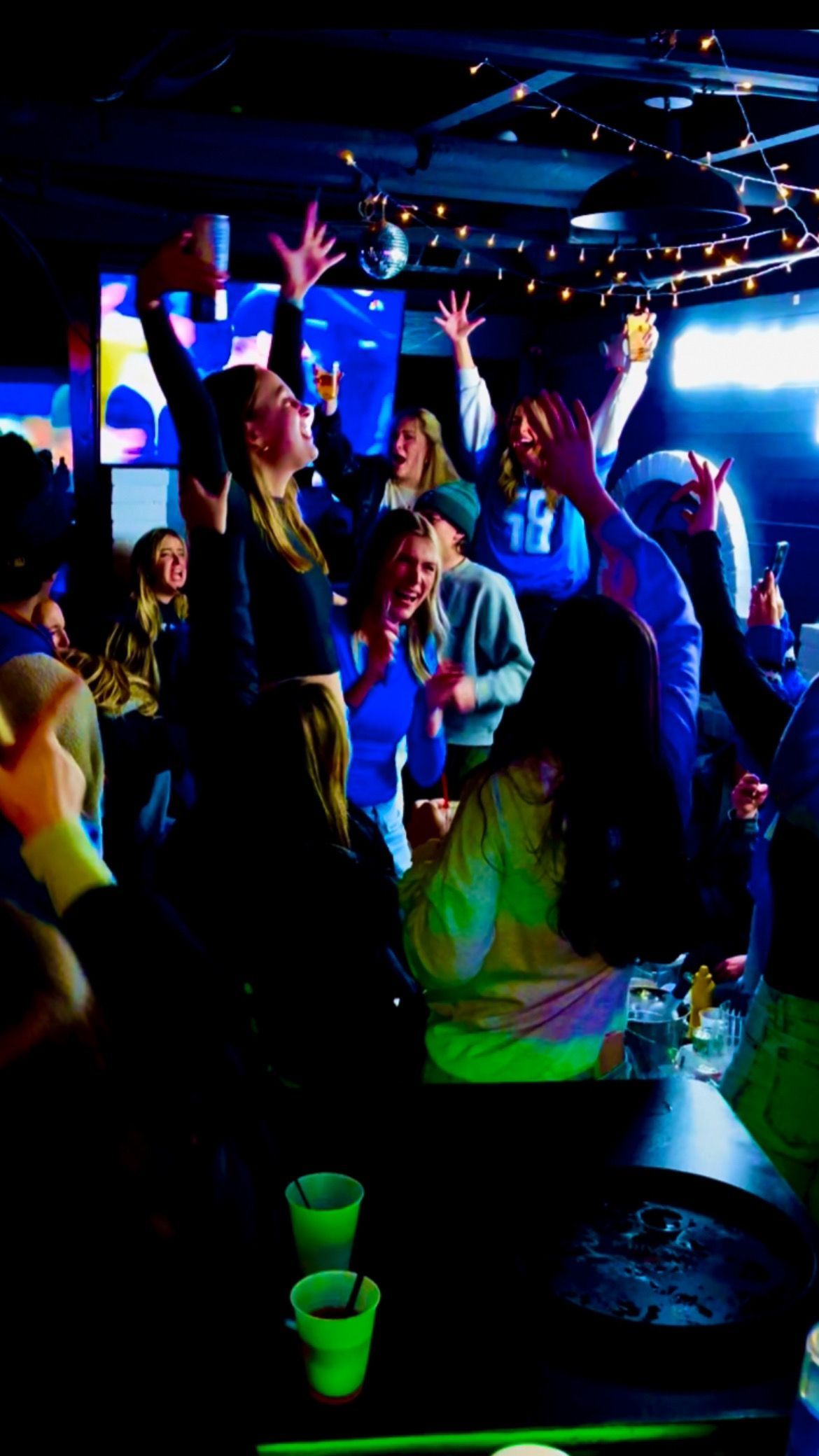 Crowded nightclub with people dancing under blue and purple lights, green cups on a table in the foreground