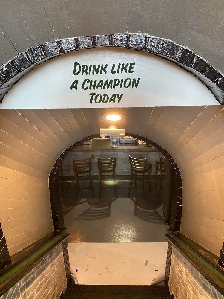 Basement stairway under a sign reading “Drink Like a Champion Today” with tables below.