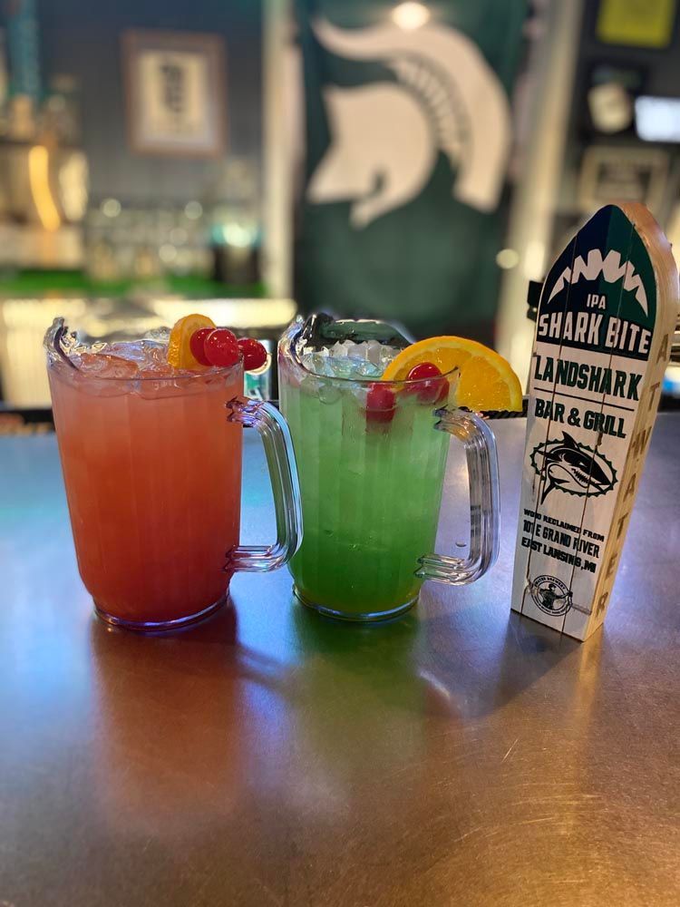 Two colorful cocktails on a bar with a garnish stick and a branded menu tent.