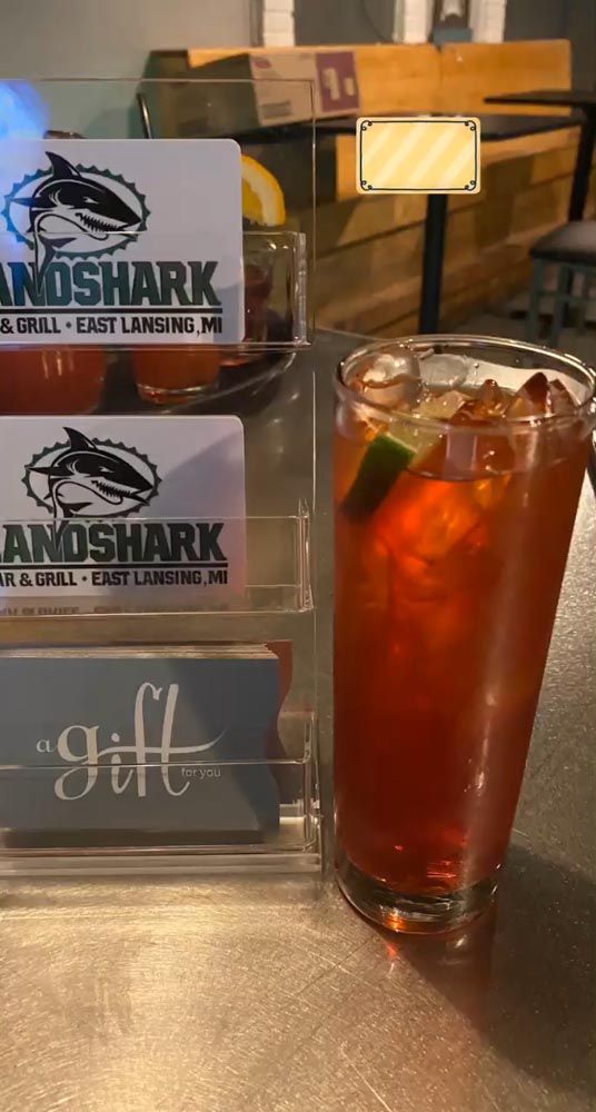 A tall iced red drink in a glass beside a table display for Landshark beer and a “Gulf” sign.