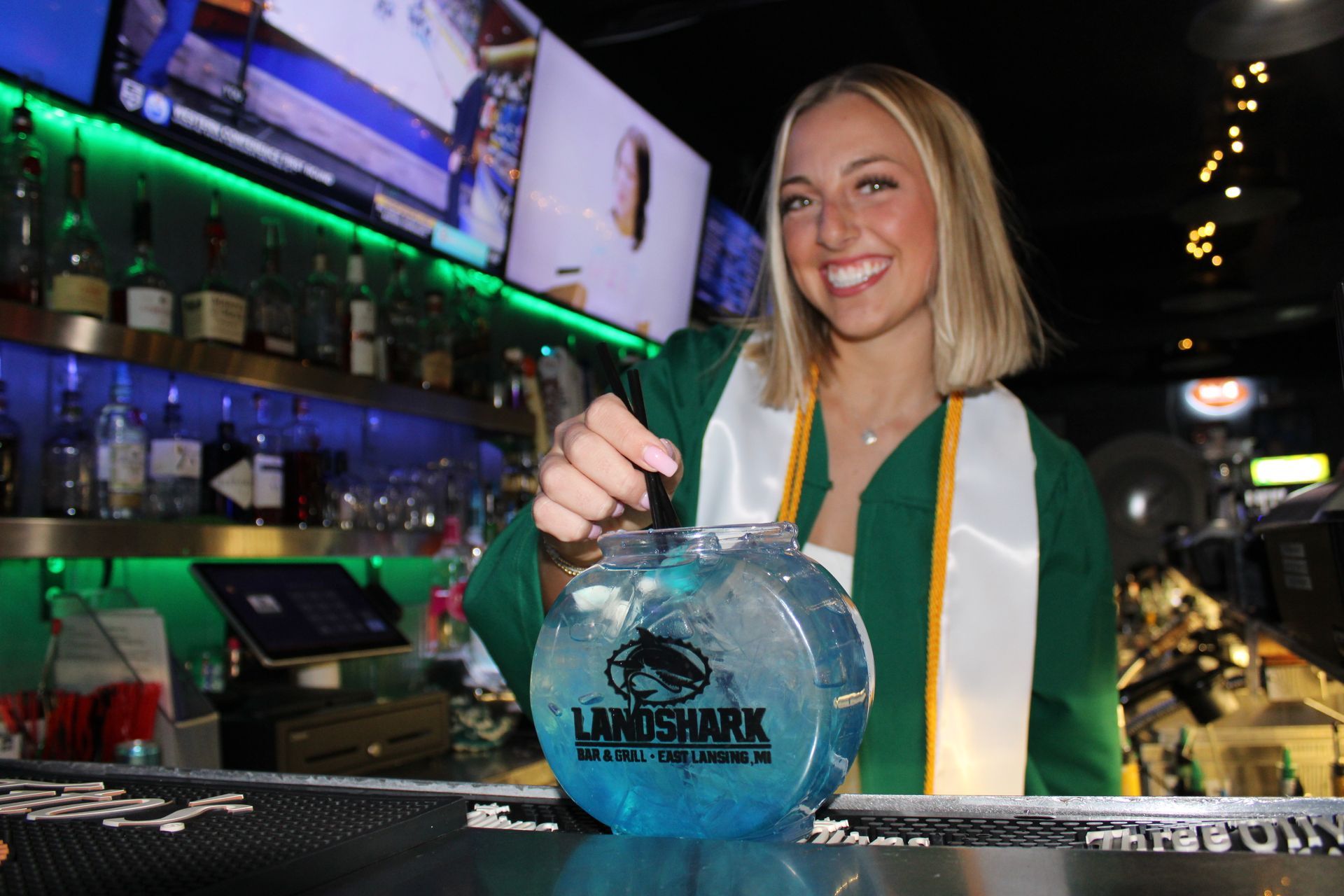 Bartender smiling behind a bar, pouring a blue drink into a LandShark glass under green lights