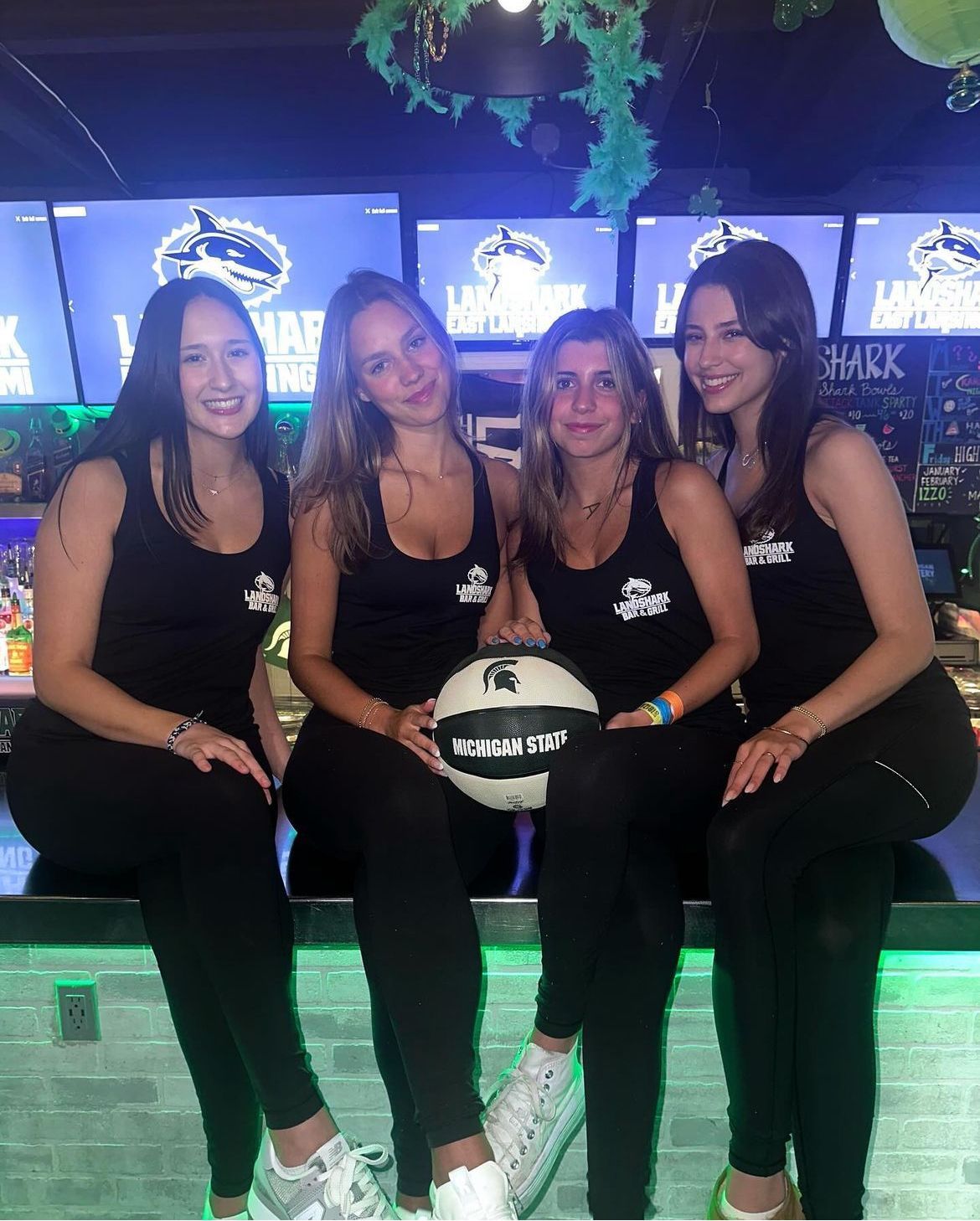 Four friends in black outfits posing at a neon-lit event booth, holding a volleyball-shaped sign.