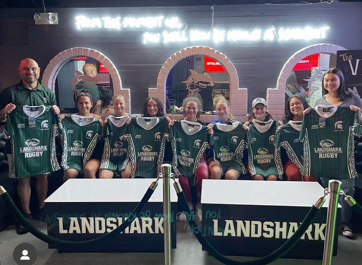Group in green sports jerseys posing with trophies behind a Landshark banner at an indoor event.