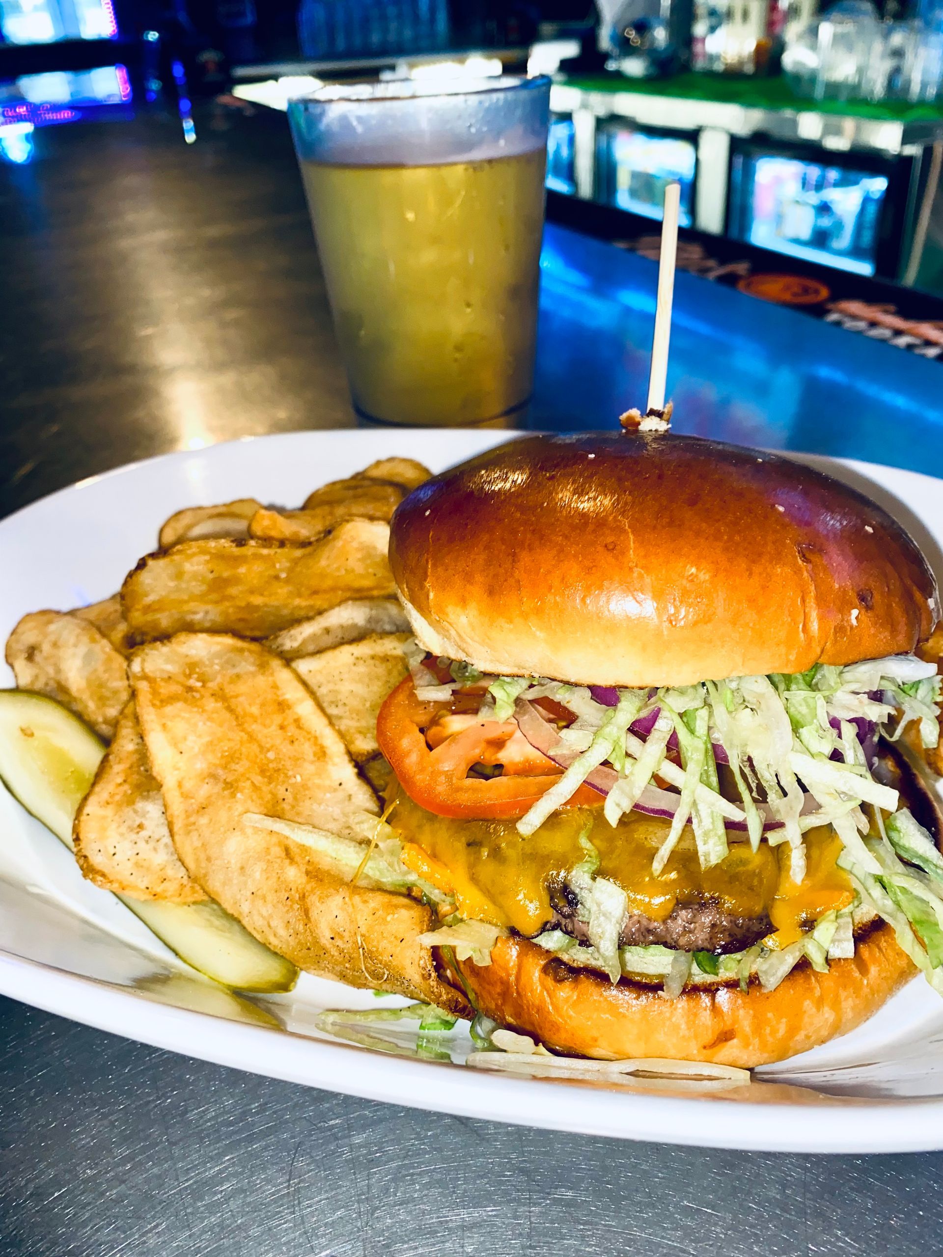 Burger with chips and pickle on a white plate at a bar, with a drink in the background