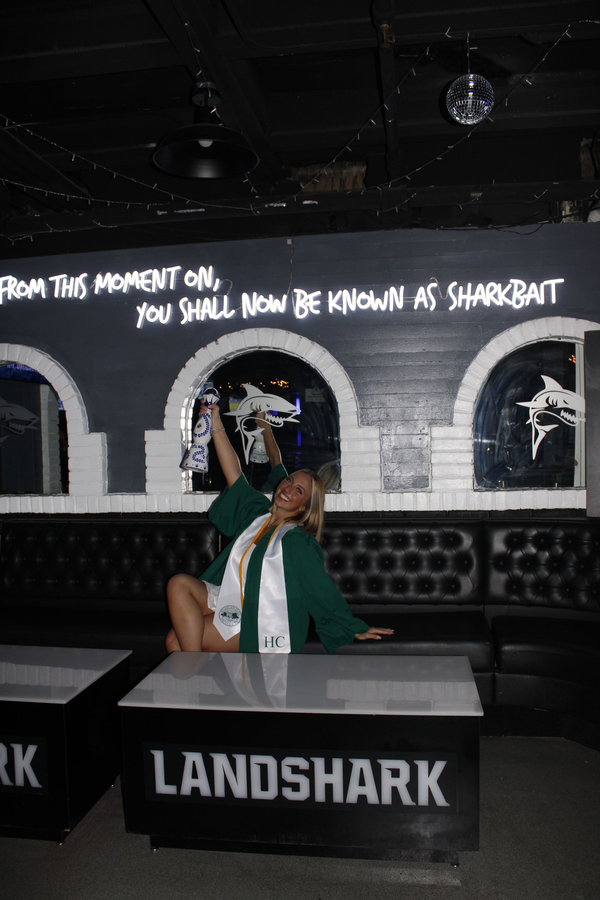 Person posing at a LANDSHARK bar beneath a neon sign that reads, “You shall now be known as Sharkbat.”