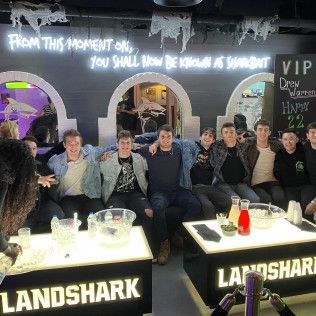 Group of friends seated at a Landshark bar table under neon signs, posing and smiling indoors.
