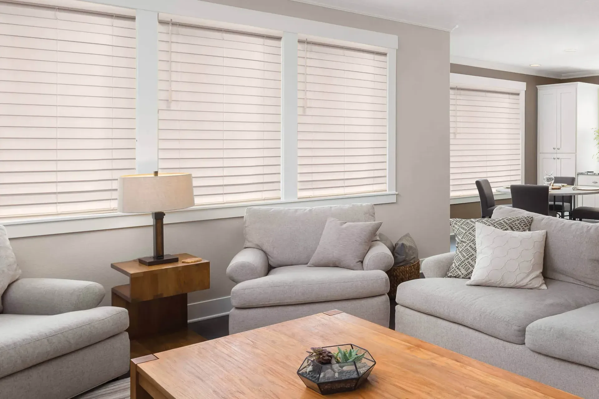 A light-filled living room featuring beige upholstered furniture, a wooden coffee table, and windows covered by white blinds.