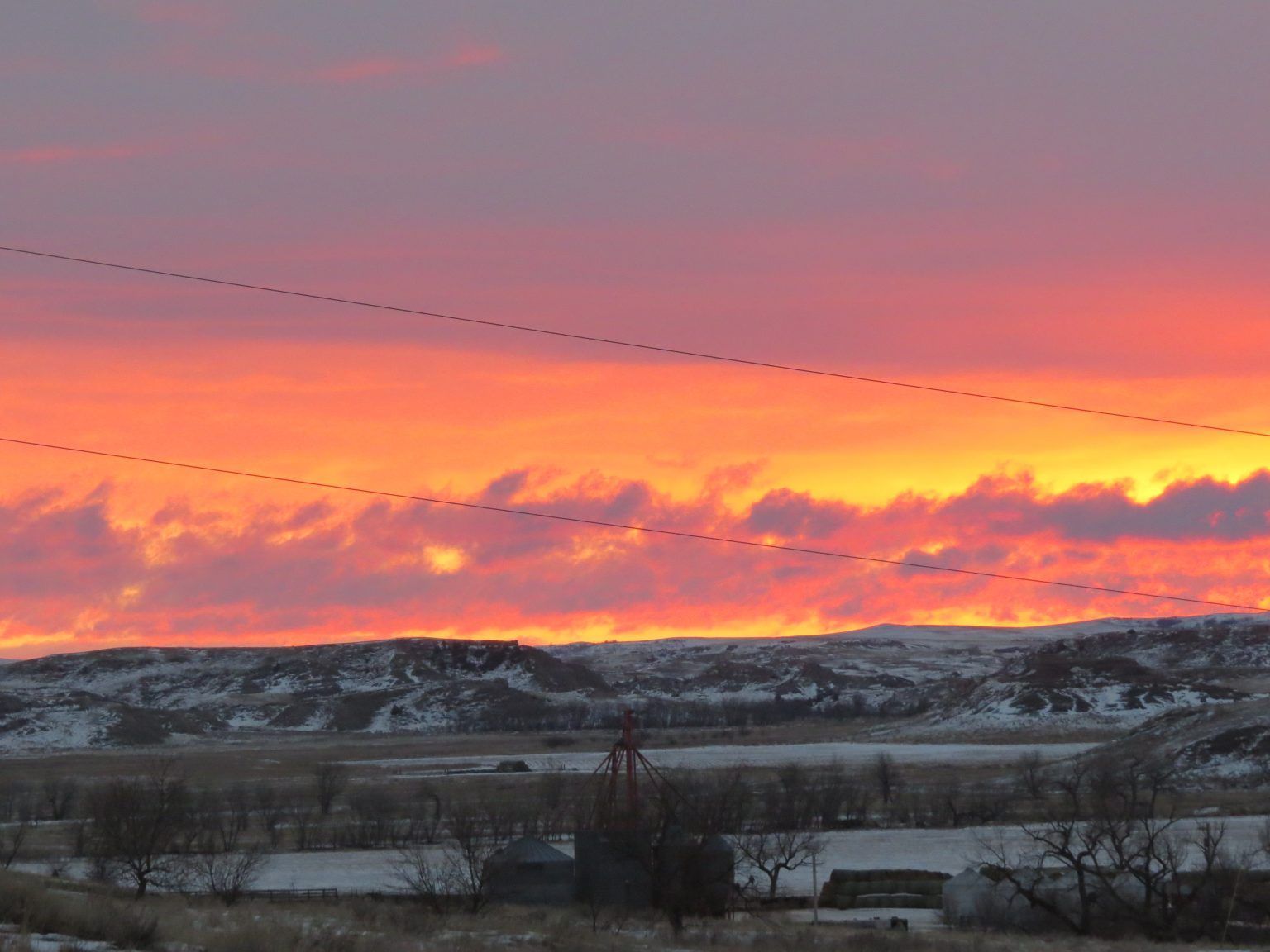 A sunset over a snowy landscape with power lines in the foreground