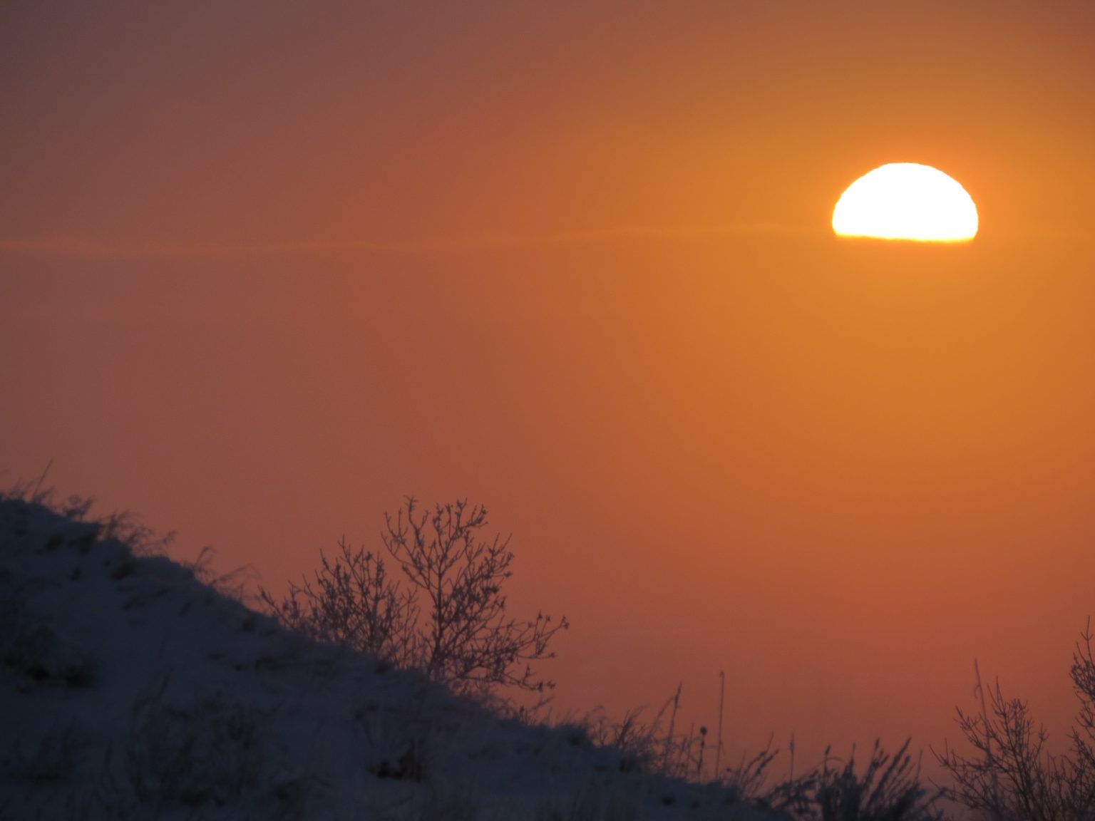 The sun is setting over a snowy hill with trees in the foreground
