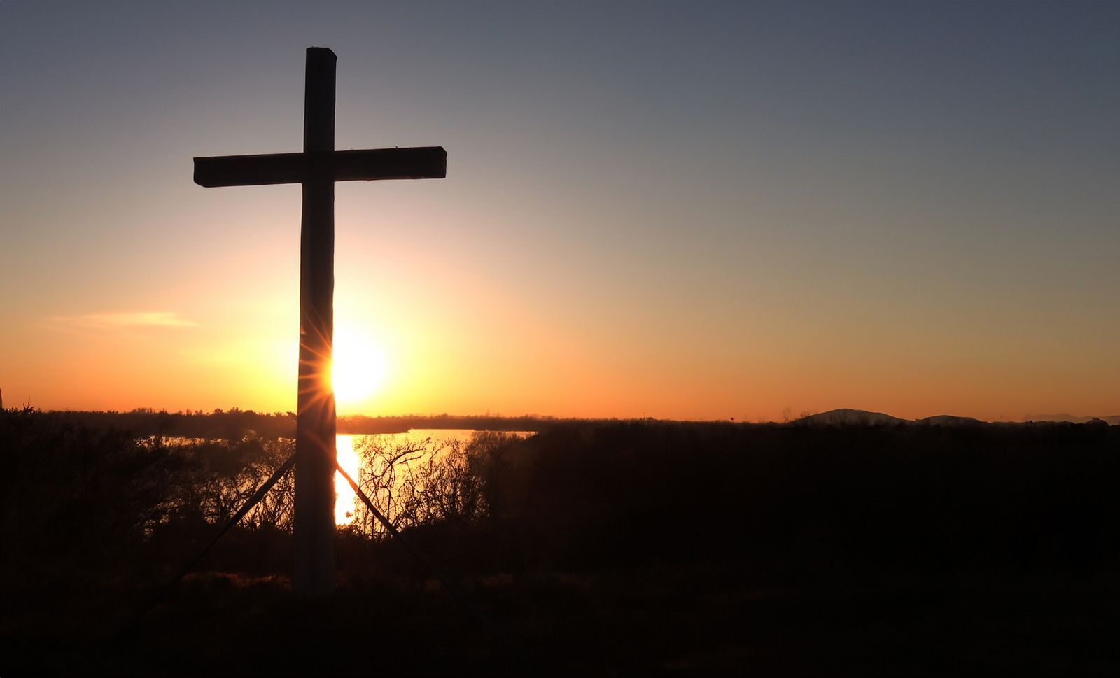A cross is silhouetted against a sunset over a body of water.