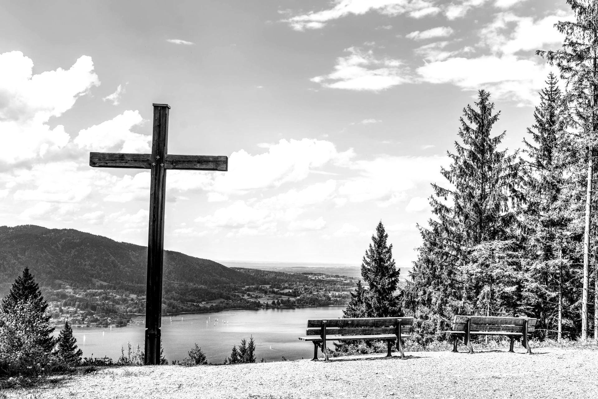 A black and white photo of a cross on top of a hill overlooking a lake.