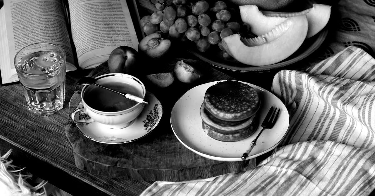 A black and white photo of a table with pancakes , fruit , a cup of coffee and a book.