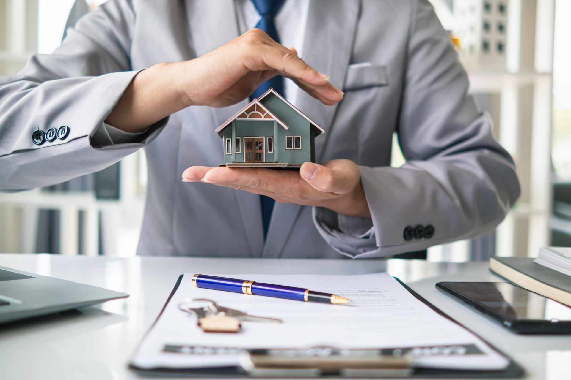 Person in suit protecting a model house with hands above paperwork, pen, and keys.