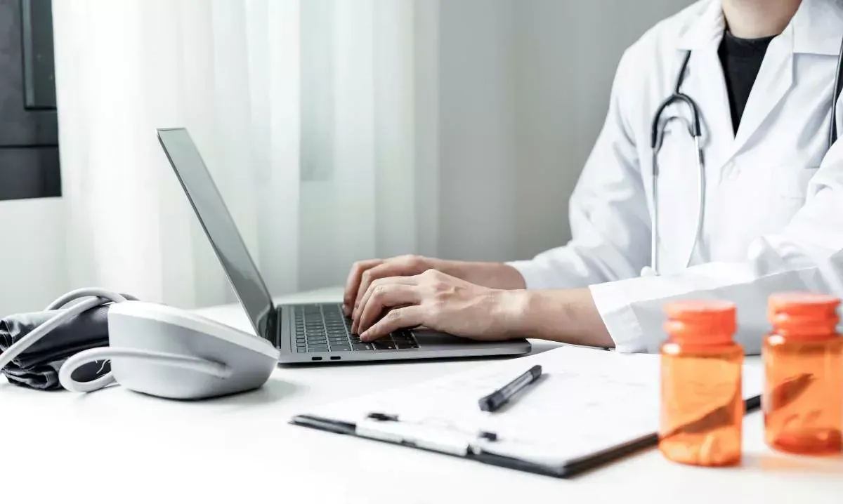 Doctor typing on a laptop at a desk with a phone, pen, and medicine bottles in a clinic