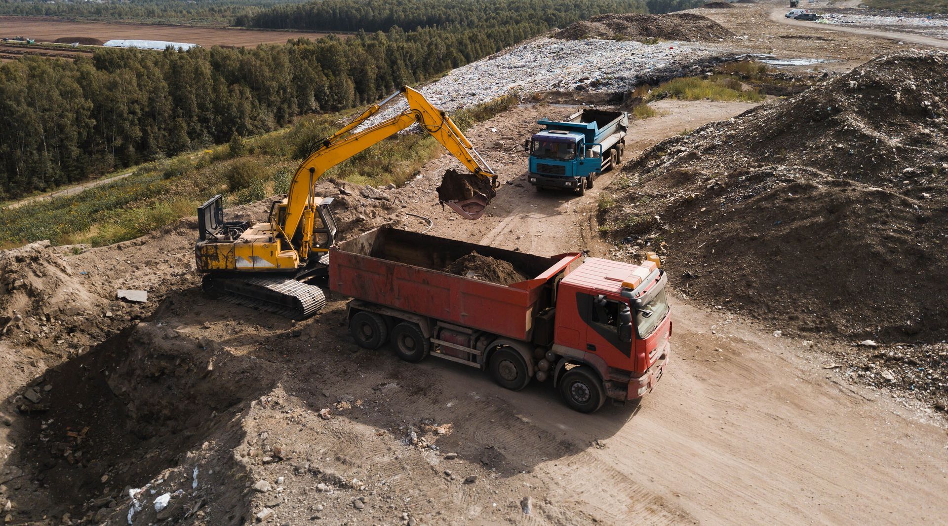 Une pelleteuse jaune charge un camion-benne rouge sur un chantier de construction ; un camion bleu se trouve à proximité.