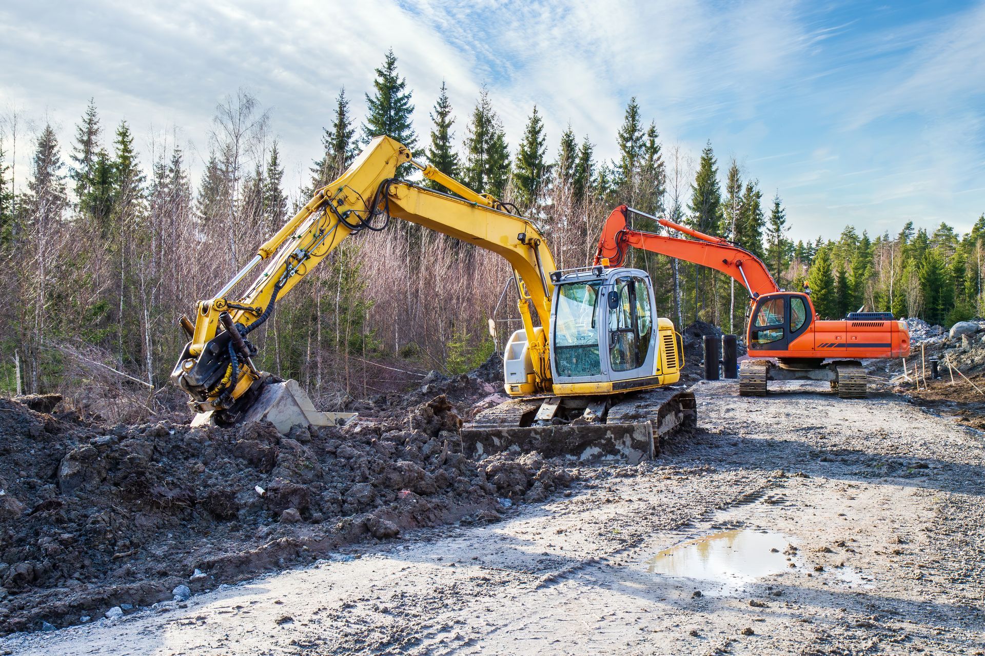 Deux excavatrices dégagent la terre et les débris d'un chantier boueux, avec des arbres en arrière-plan.