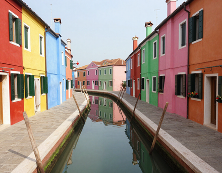 Lungo uno stretto canale, le cui acque tranquille riflettono gli edifici, si affacciano case luminose e colorate.