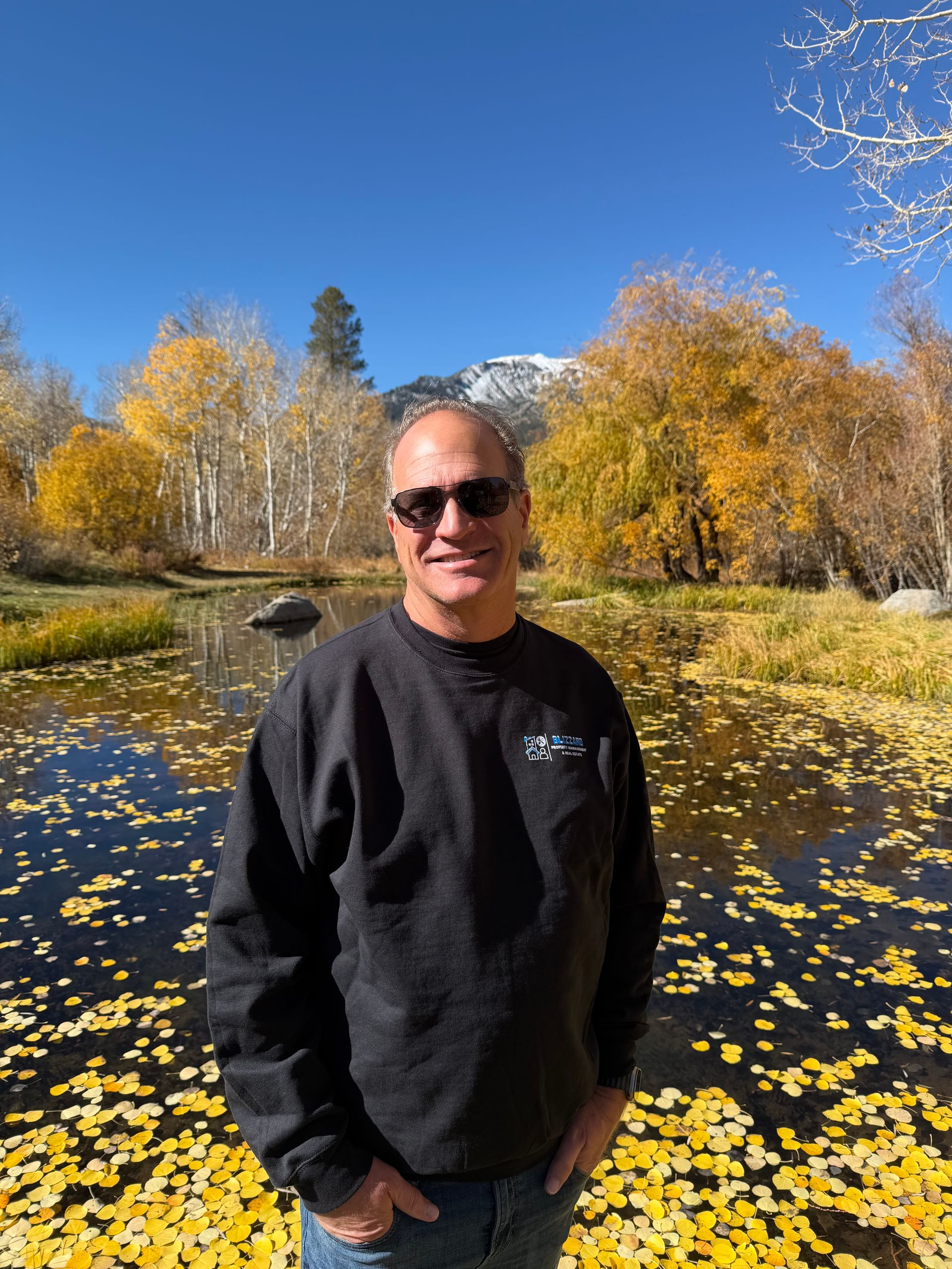 Man smiling in sunglasses beside a flower-covered pond with autumn trees and a blue sky