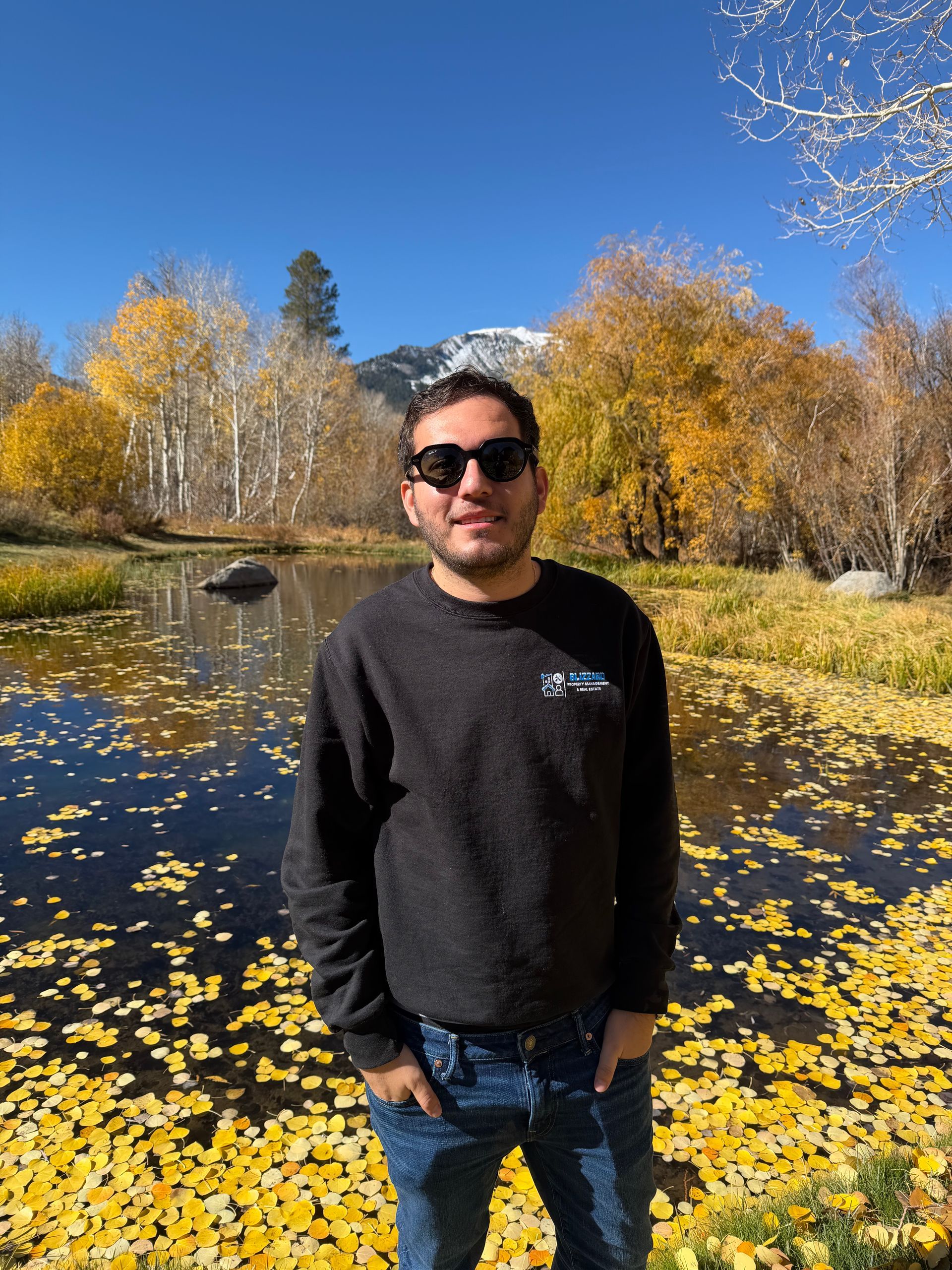 Man standing by a leaf-covered pond in autumn, wearing sunglasses and a dark sweater under a blue sky