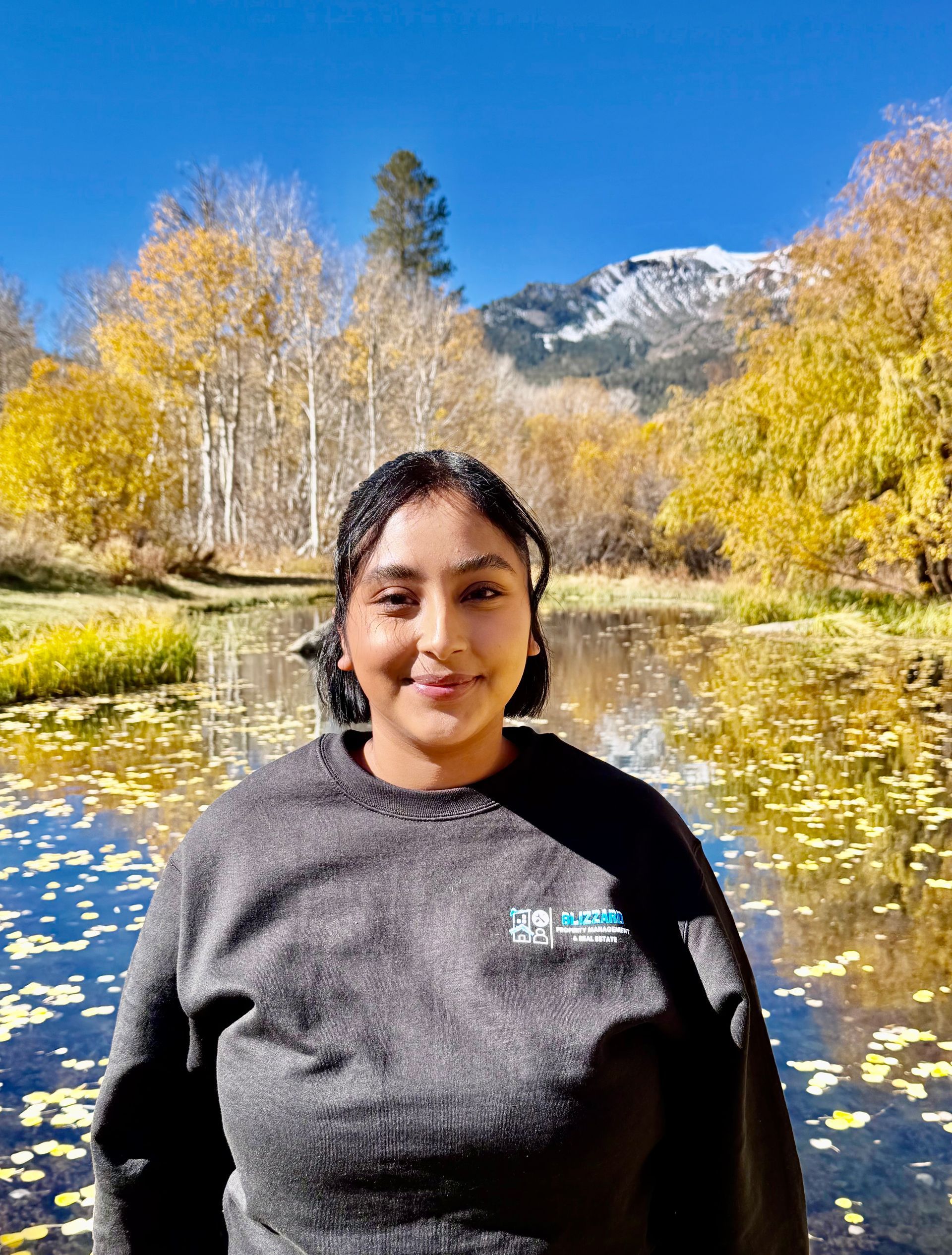 Person smiling by a river with yellow autumn trees and snowy mountains in the background