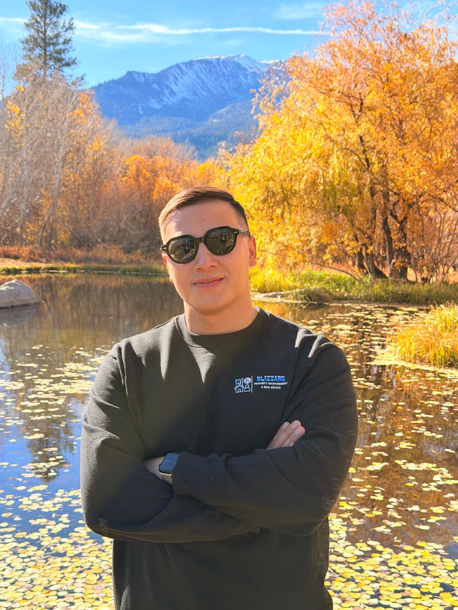 Person in sunglasses standing by a reflective lake with autumn trees and mountains in the background