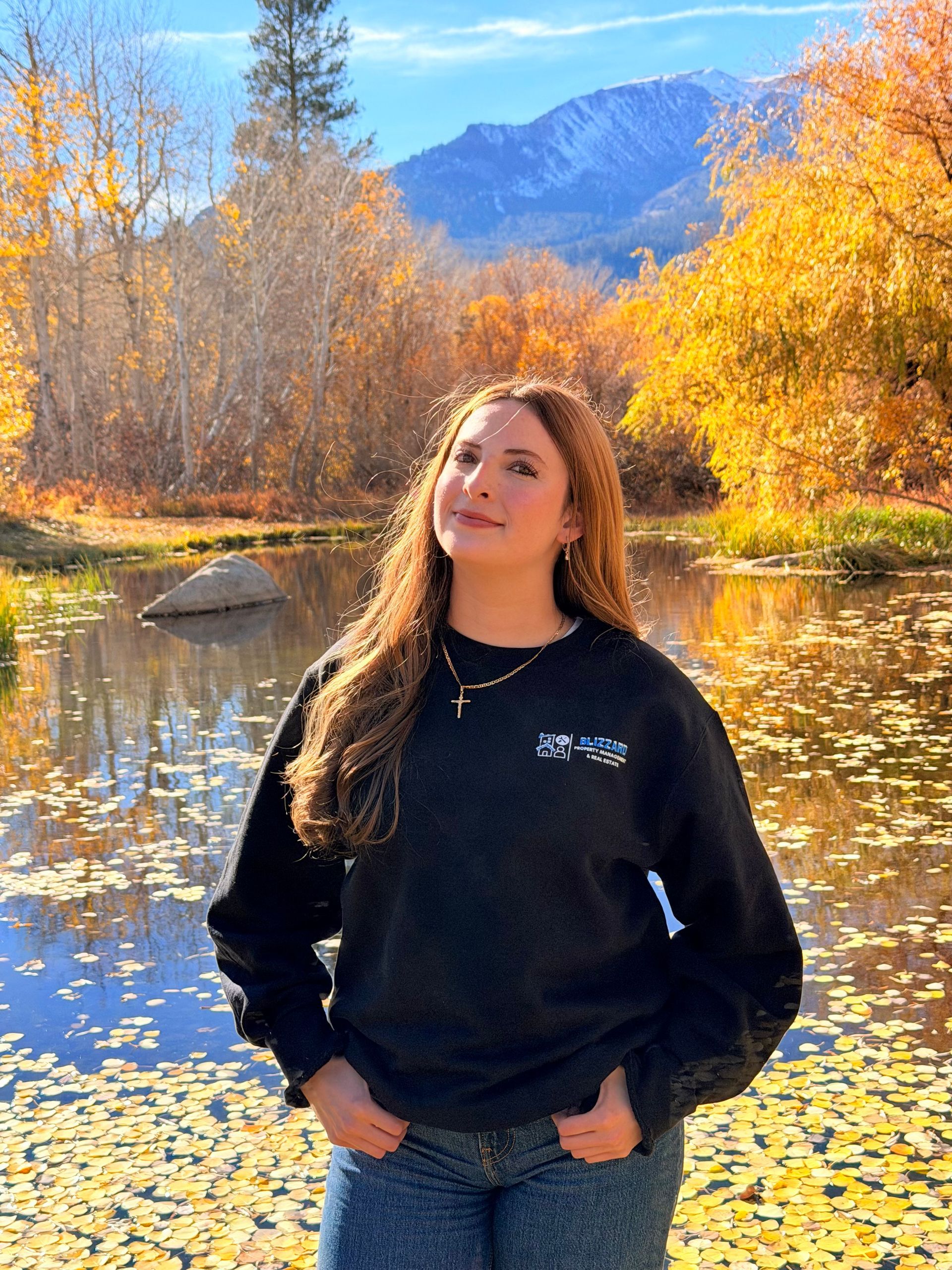 Woman standing by a reflective river in autumn, with golden trees and mountains in the background.