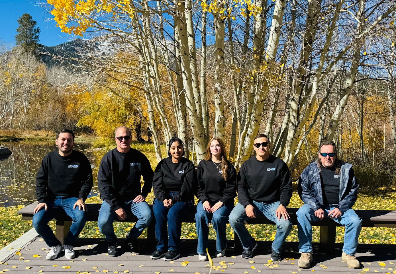 Six people sitting on a bench by a lake, with autumn trees and mountains in the background.