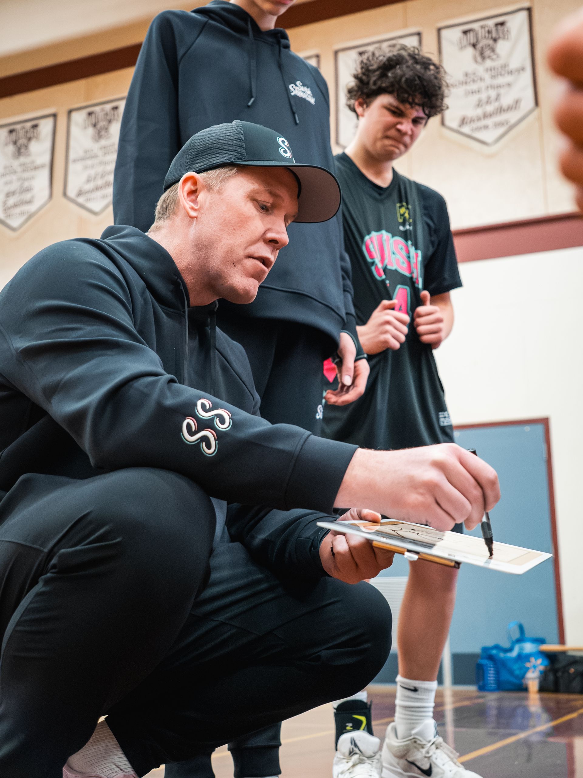 Coach kneels beside a student, demonstrating a hand drill in a gymnasium.