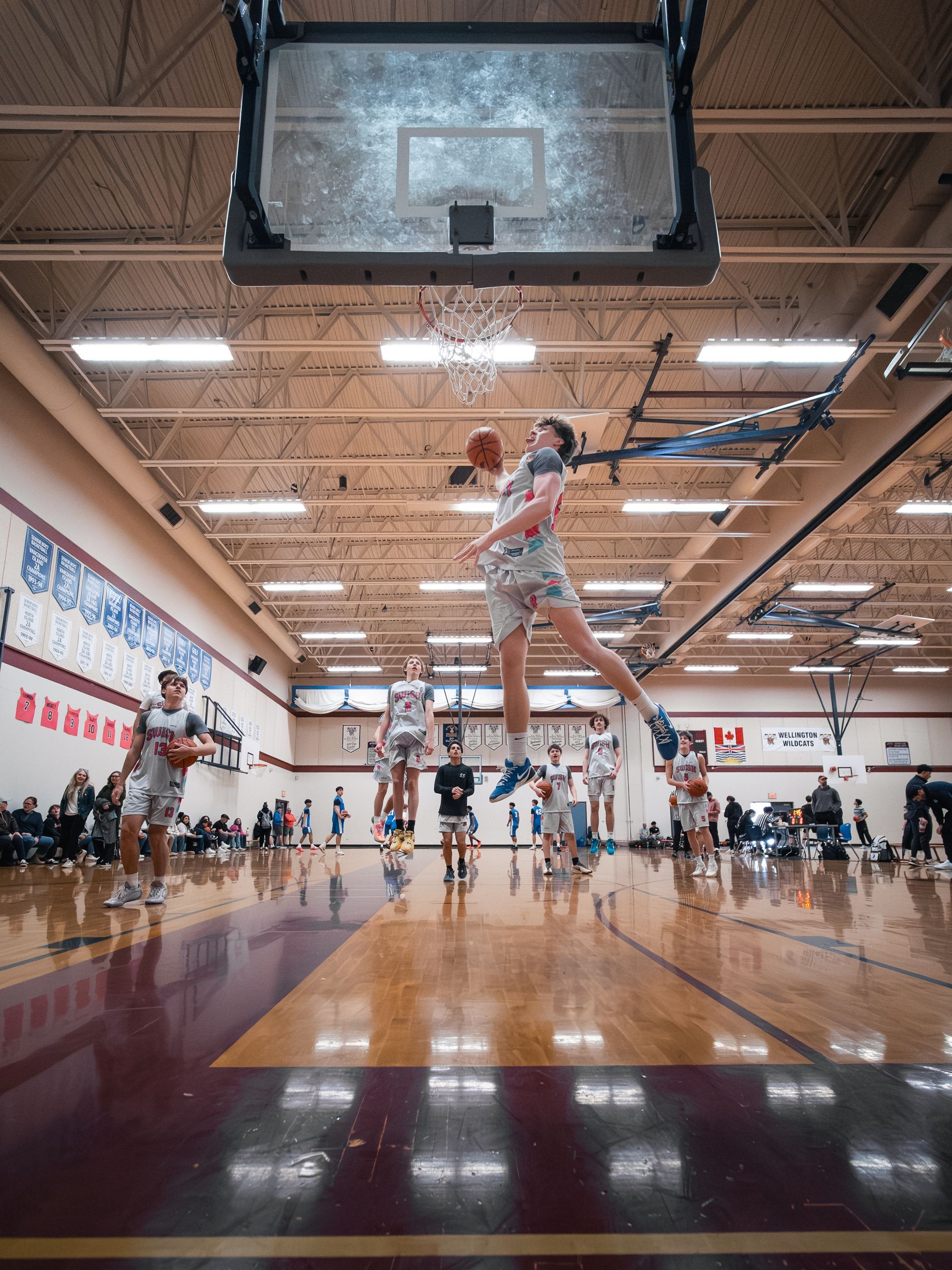 Indoor basketball game with a player midair for a layup near the hoop on a polished gym court.