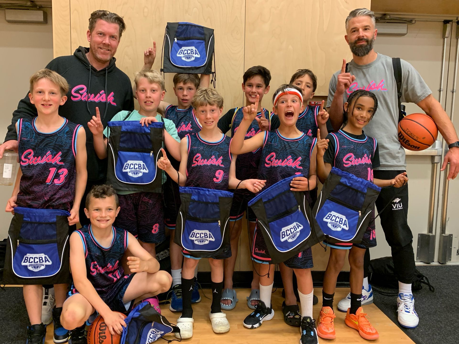Youth basketball team posing with coaches, holding blue bags in a gym