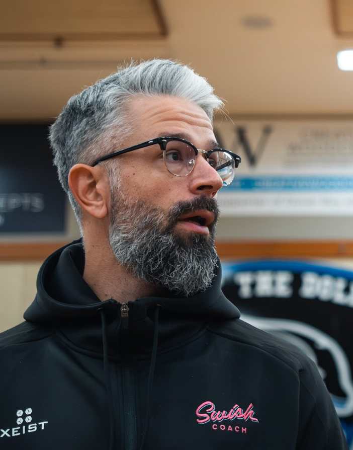 Man with gray beard and glasses in a black hoodie, looking left indoors.