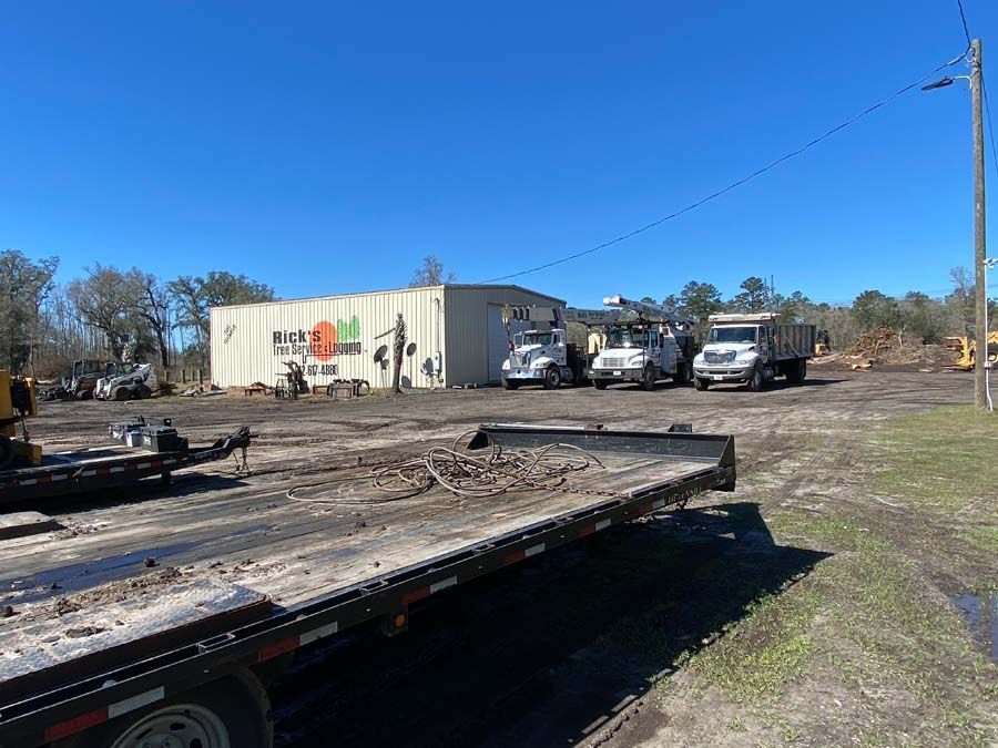 A flatbed trailer sits in the foreground of a dirt lot with a warehouse and several large trucks under a clear blue sky.
