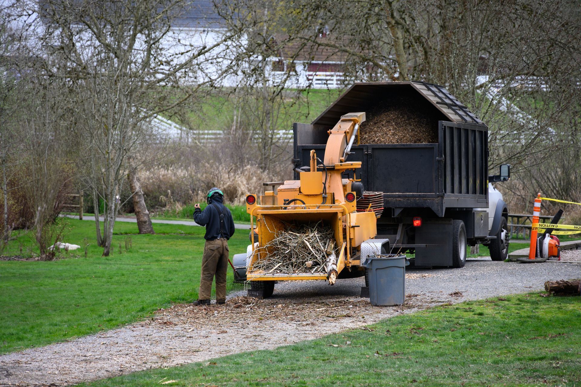 A person operates a yellow wood chipper, feeding debris into the machine while chips collect in a truck bed nearby.