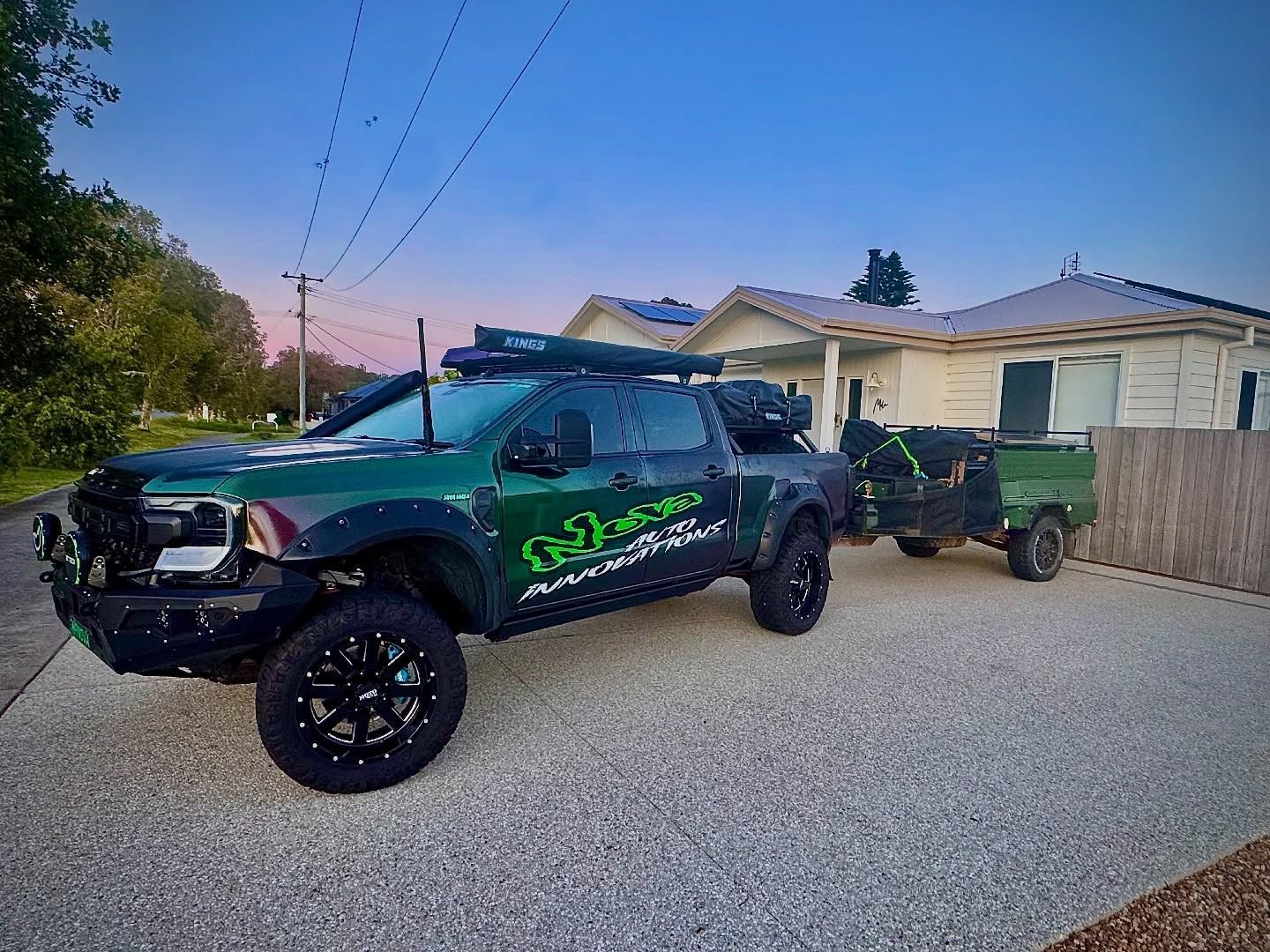 A dark green, modified pickup truck with large tires and decals, parked in a residential driveway while towing a trailer — Nova Auto Innovations in Charlestown, NSW