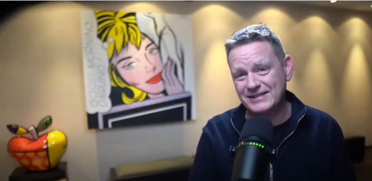 Um homem está em pé diante de um microfone em frente a uma pintura de uma mulher.