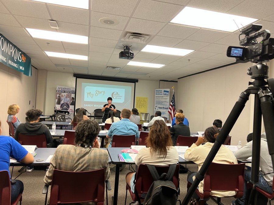 A presenter leads a class in a room. Students sit at desks, a projector displays a slide, and a camera records.