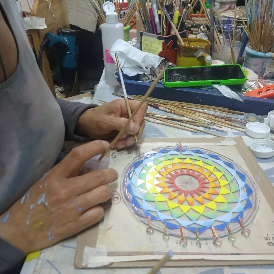 Un artista pinta un colorido mandala sobre una tabla redonda en un banco de trabajo con pinceles y materiales a su alcance.