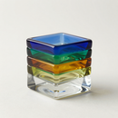 Vasos cuadrados de vidrio apilados en azul, verde, ámbar y transparente sobre un fondo claro.