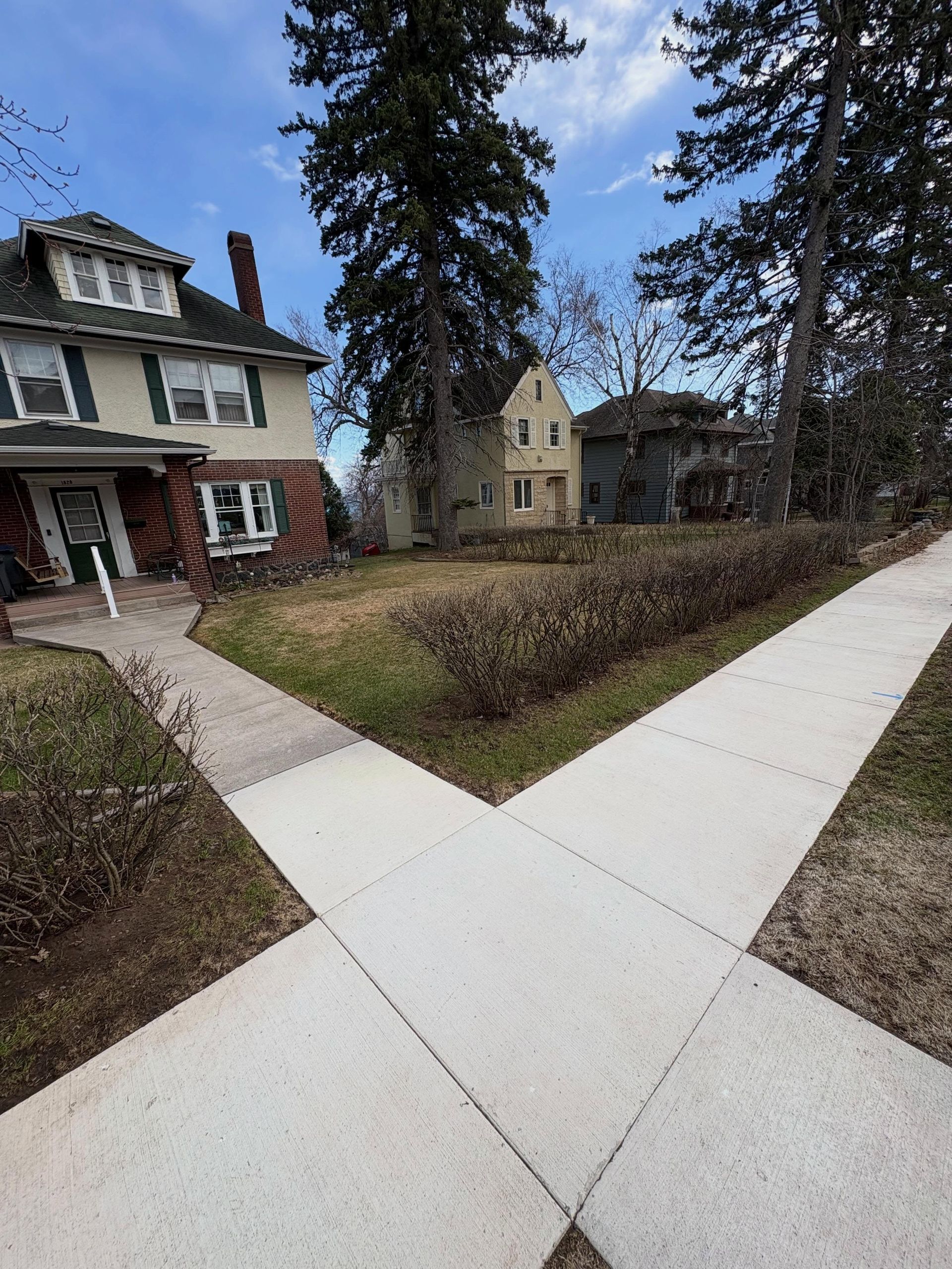 Sidewalks crossing a grassy residential yard beside two houses and tall trees on a cloudy day