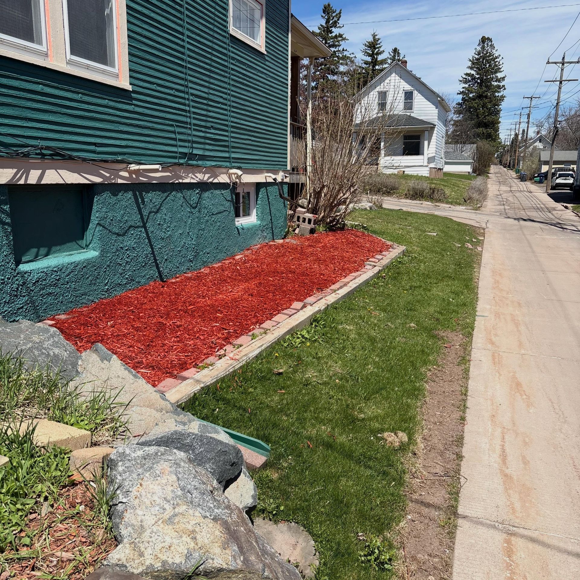Sidewalk beside a green house with red mulch, grass strip, and a residential street in the background