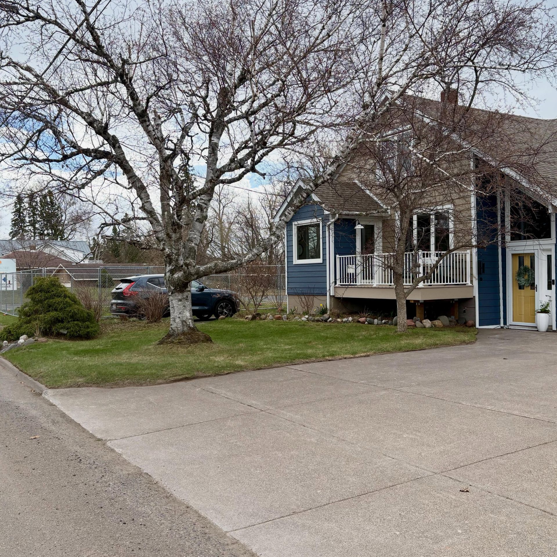 Blue house with a porch beside a leafless tree and parked cars on a quiet street.