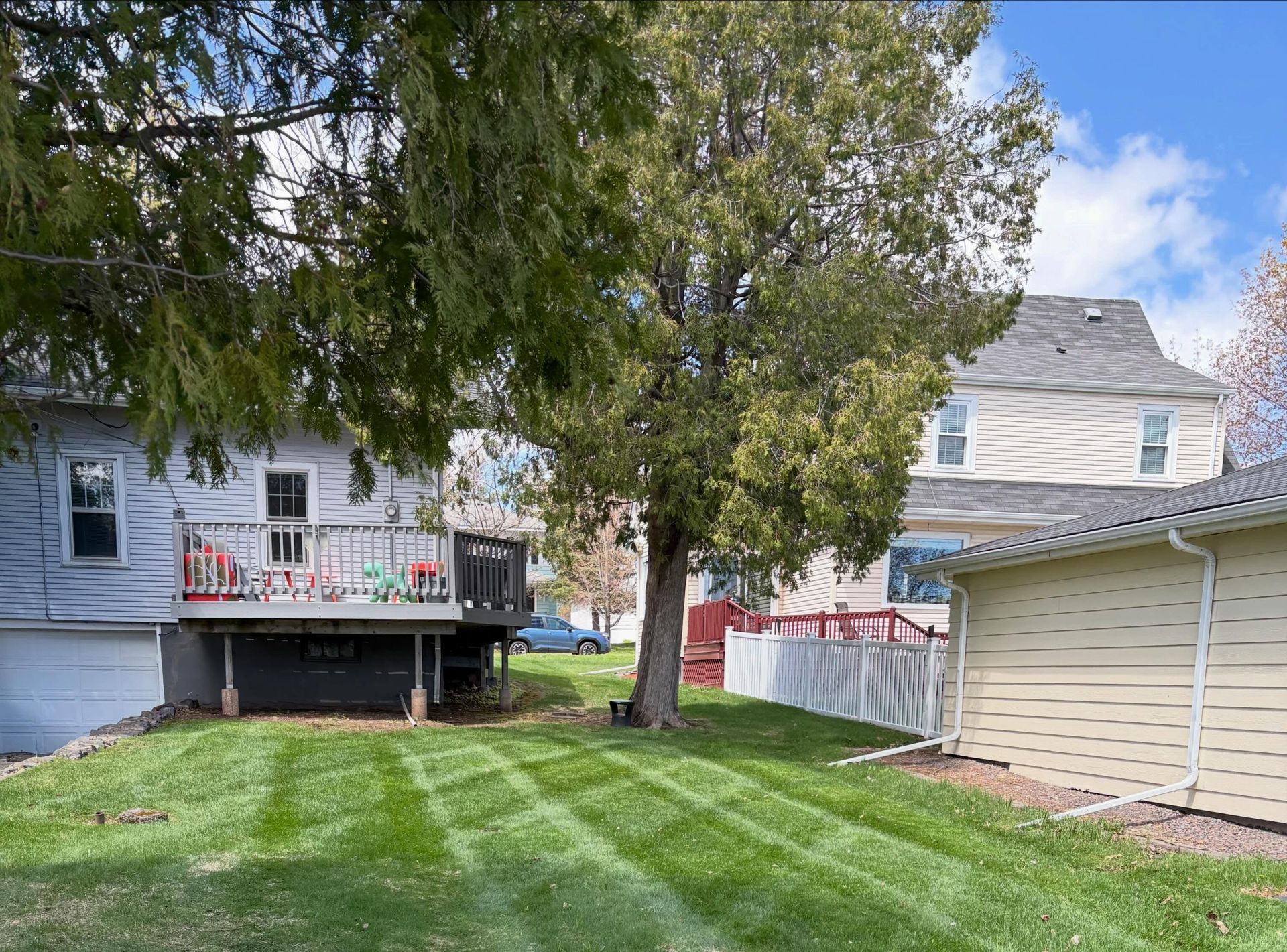 Backyard with green lawn, large trees, a white house deck, and beige garage on a sunny day
