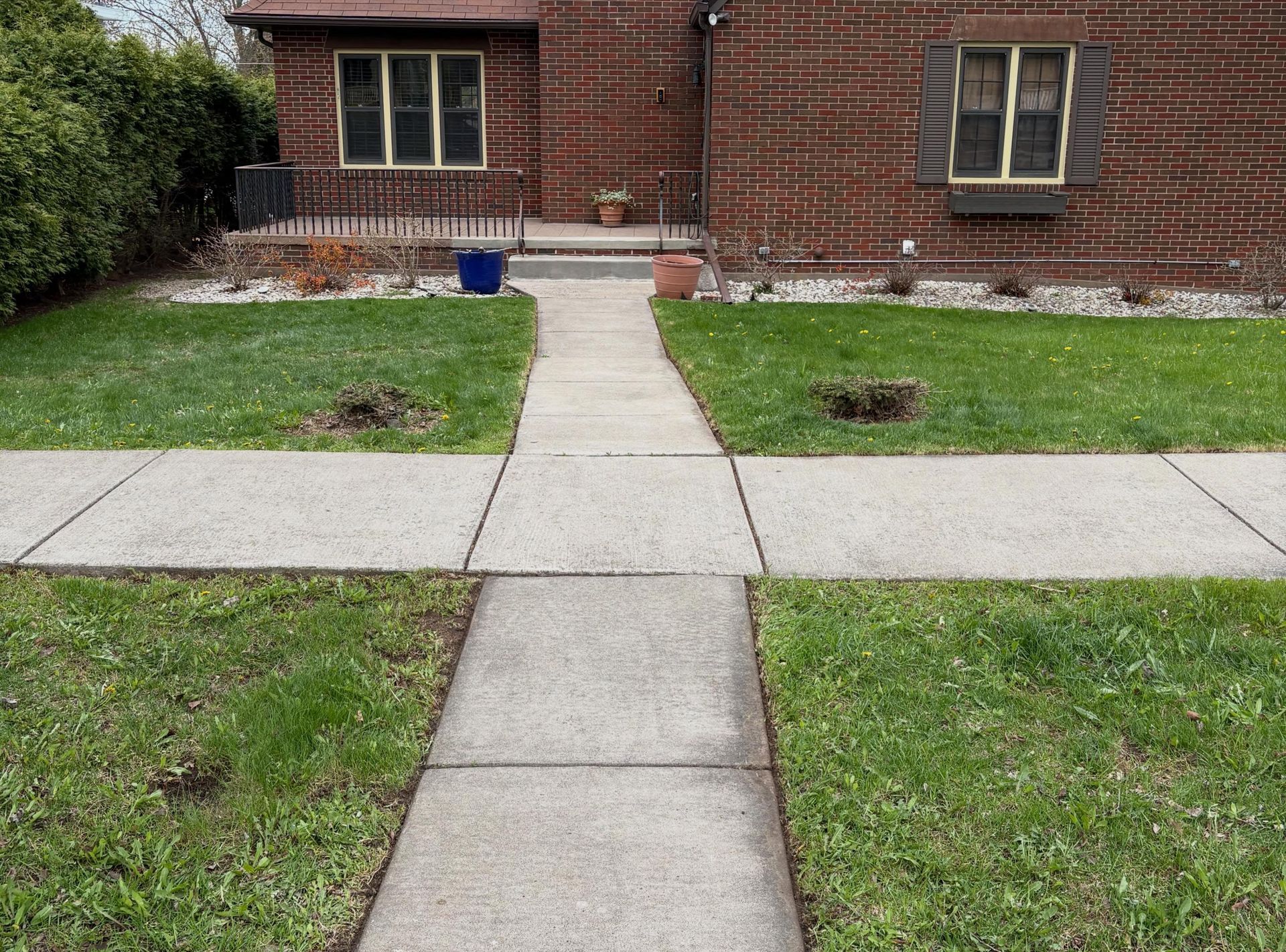 Concrete walkway leading to a brick house porch, with grass lawns and two small garden beds.