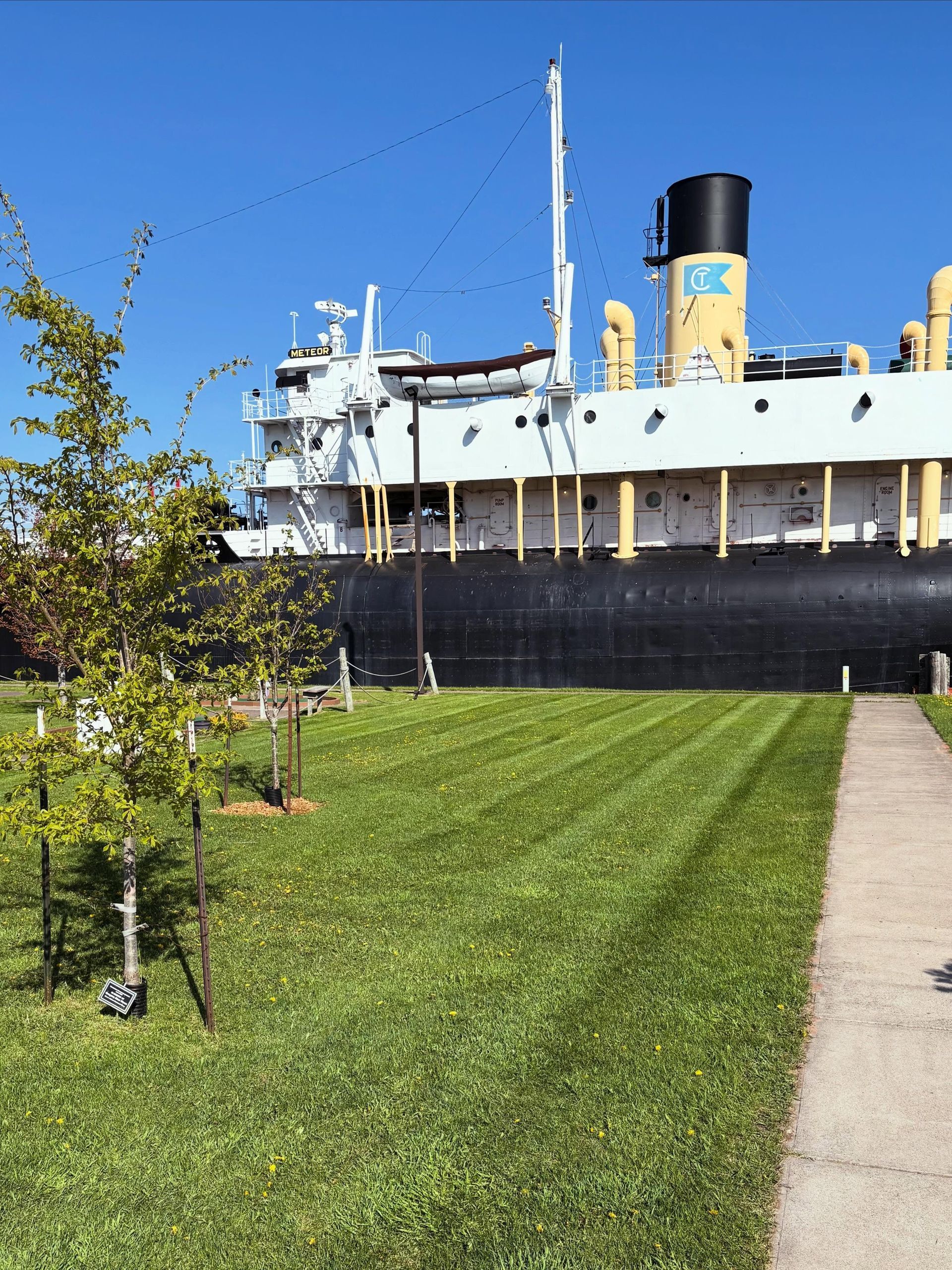 Large white ship with a black funnel behind a green lawn and sidewalk on a sunny day