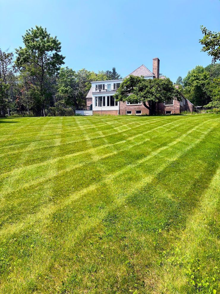 Large brick house with white upper floors beside a striped green lawn and trees under a blue sky