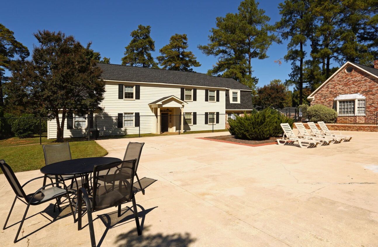 A two-story white building with black shutters and a red brick annex, with a concrete patio featuring outdoor furniture.