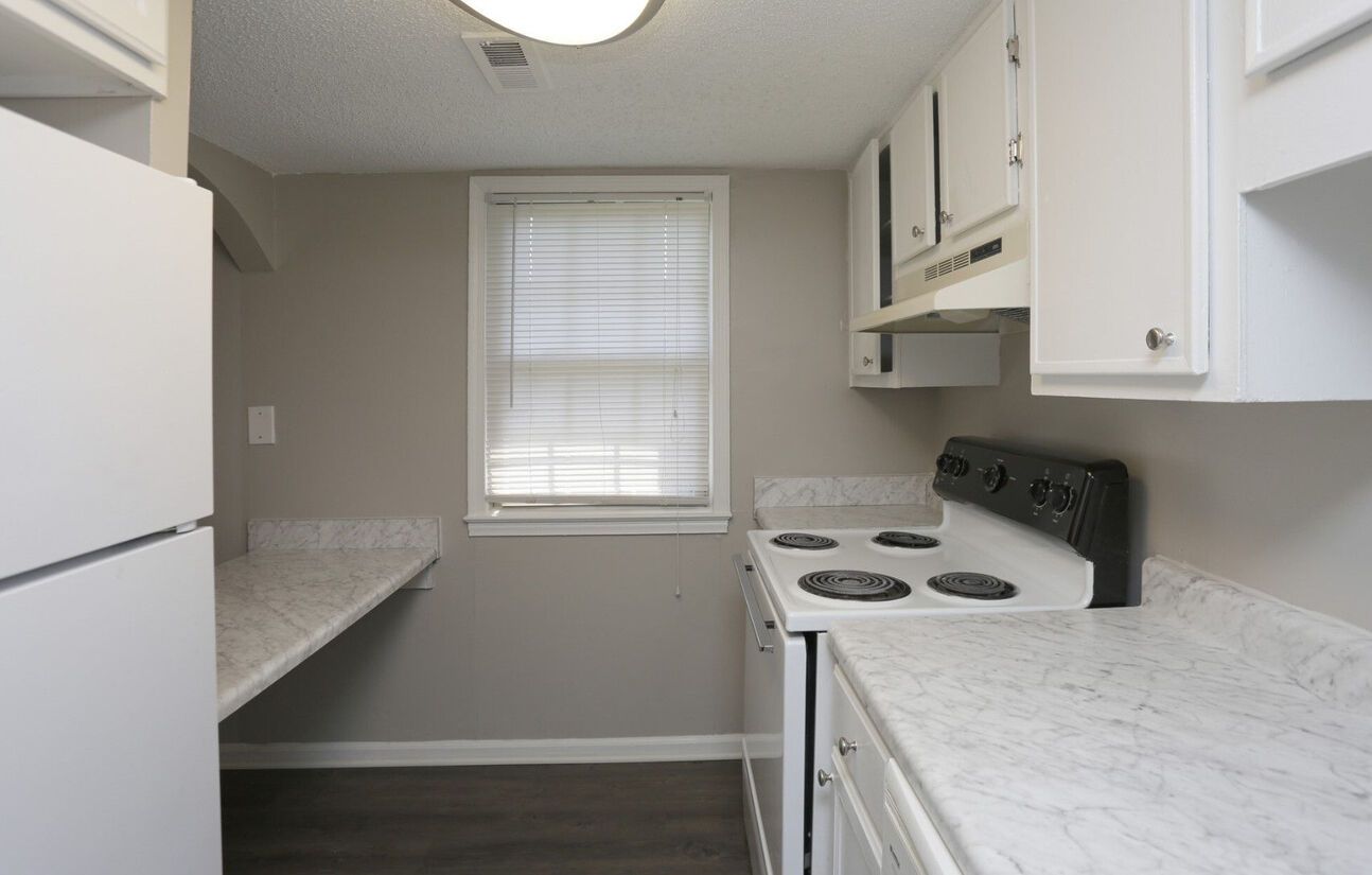 A small kitchen with white cabinets and appliances, gray walls, and a window with blinds.